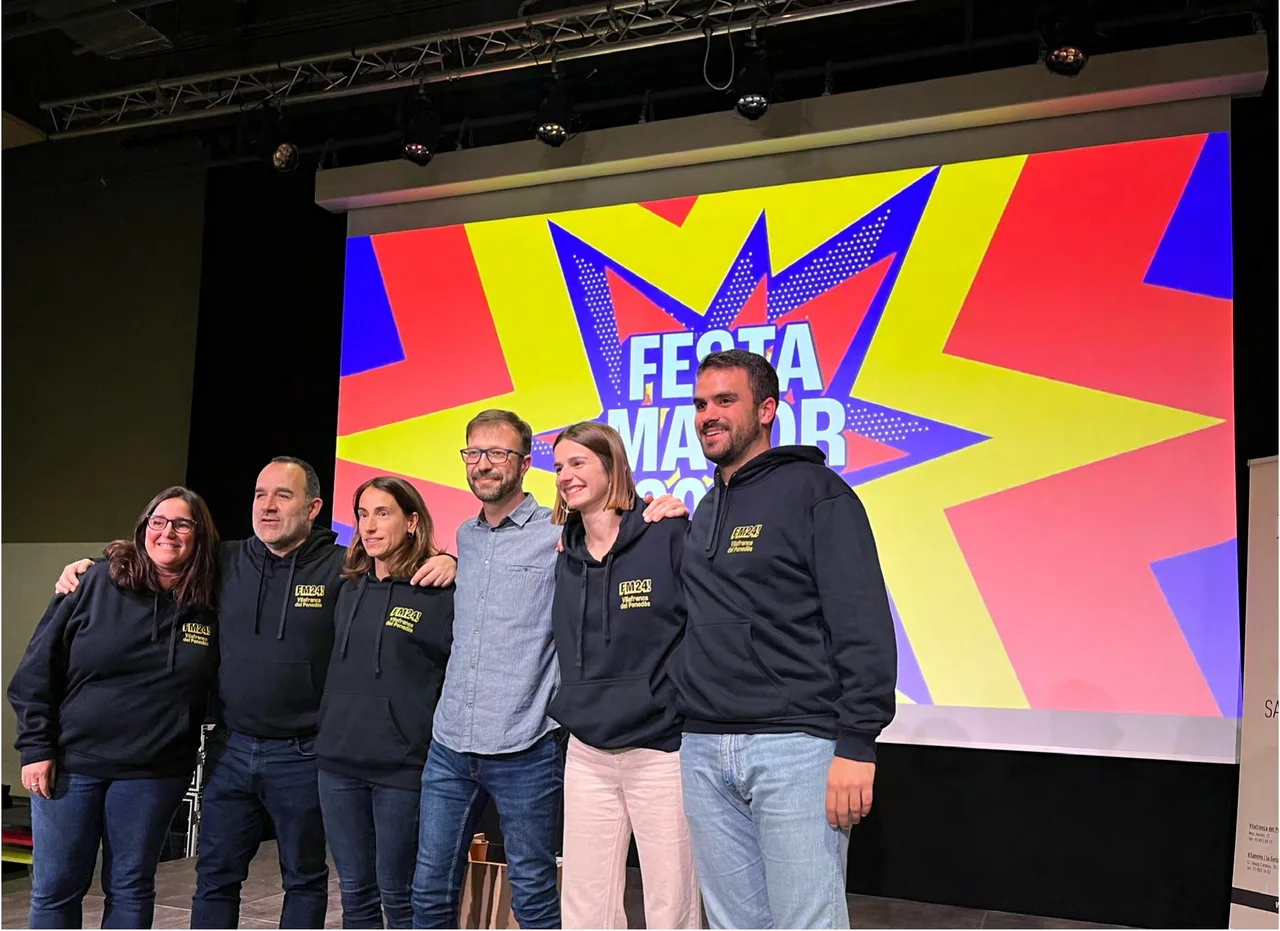 Equip de la Festa Major davant la pantalla de l'escenari amb la imatge explosiva en vermell, groc i blau