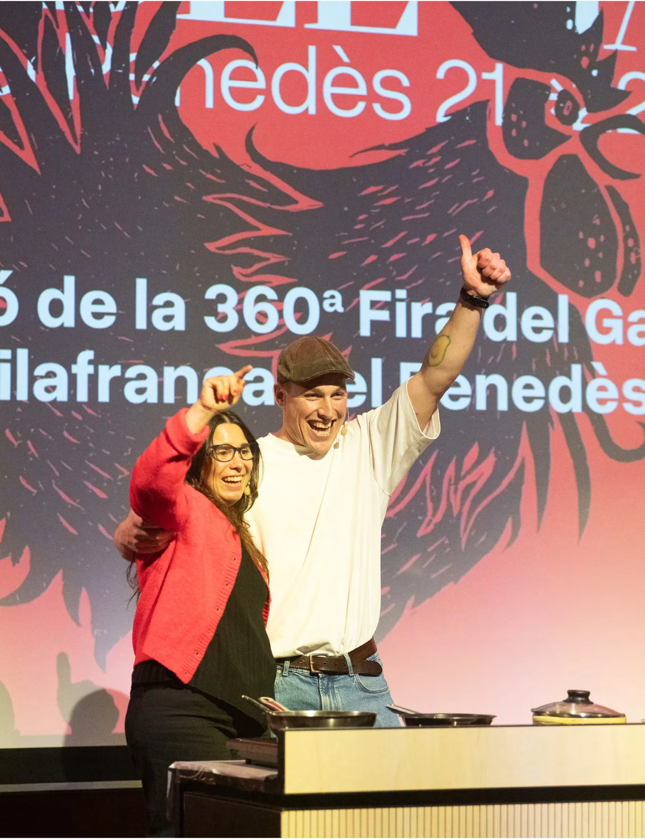 Presenters on stage at the 360th Fira del Gall in front of a red graphic backdrop